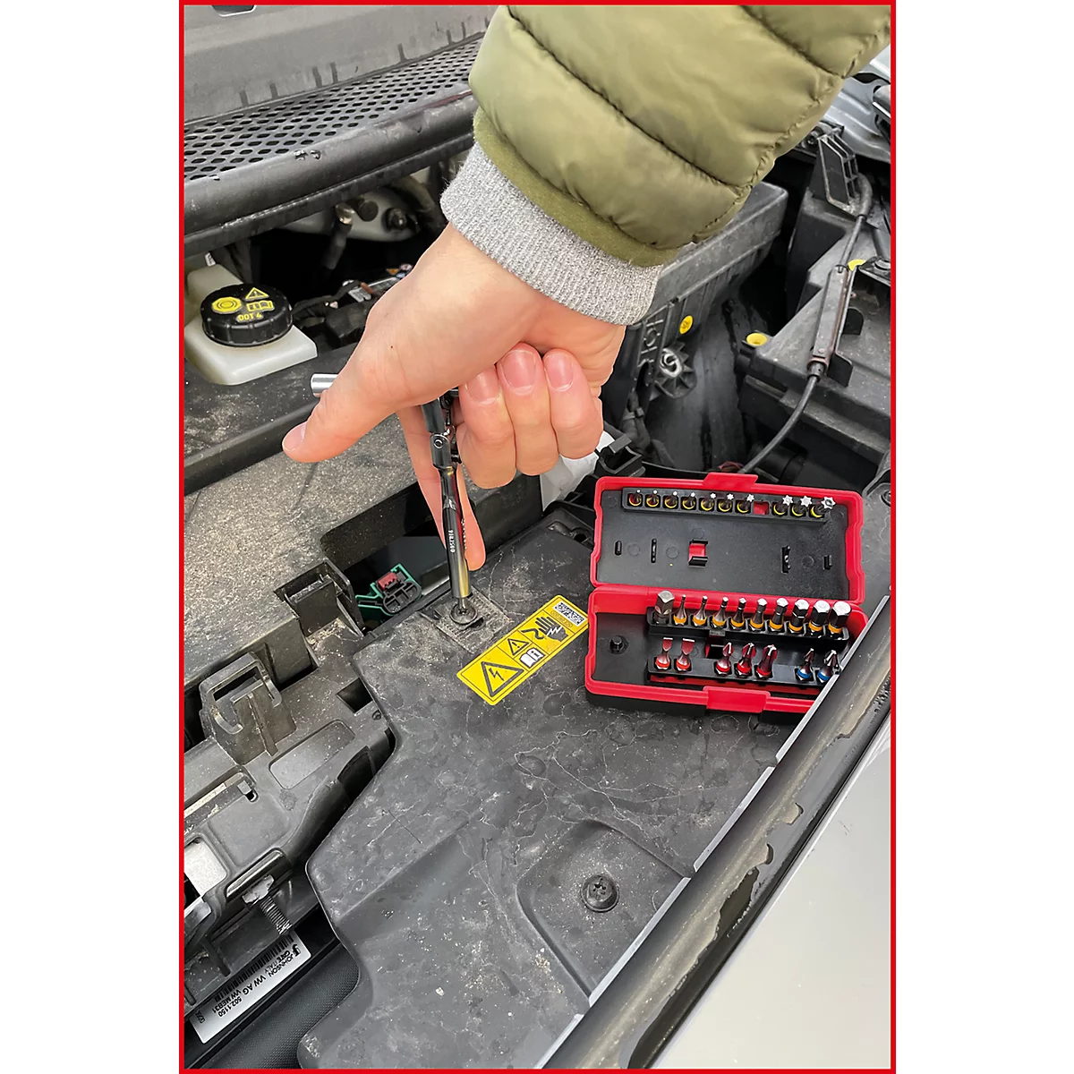 Person schraubt mit Schraubenzieher an einem Auto. Roter Werkzeugkasten mit Bits daneben. Gelbes Warnschild.